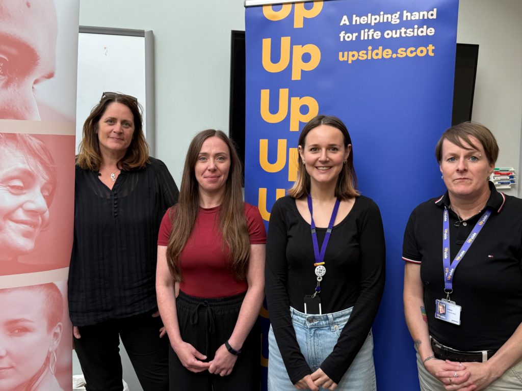 Four women standing in front of promotional banners at the launch of the Upside service in Moray. One banner displays the Upside logo and slogan "A helping hand for life outside" with the website upside.scot