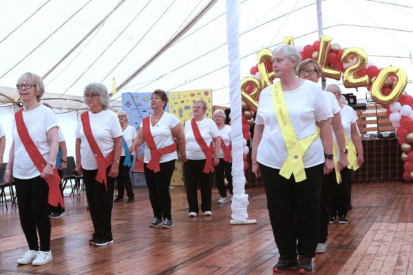Two columns of women in white t-shirts, black trousers and trainers, are wearing red or gold sashes and are about to perform a marching routine in the giant marquee.