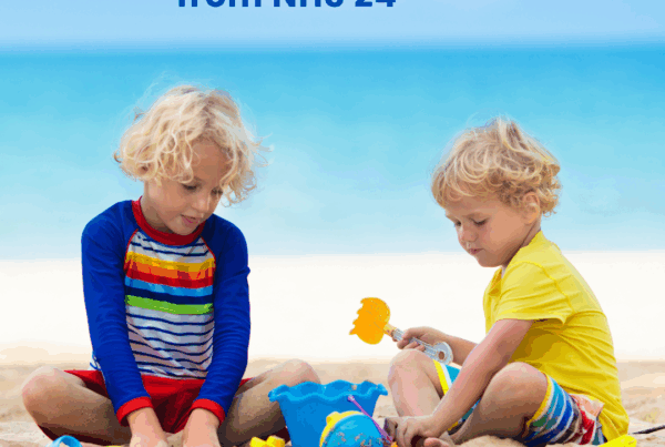 Two young children in shorts and T-shirts sit playing on the golden sand of a beach as the blue sea sparkles behind them.