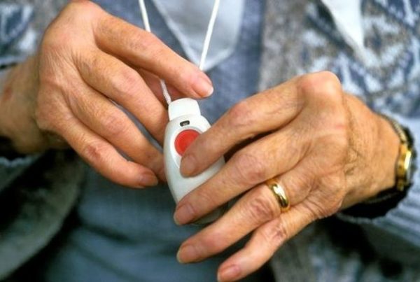The hands of an older woman clasp the community alarm button which she is wearing a a pendant around her neck.