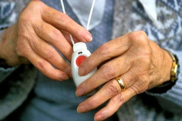 The hands of an older woman clasp the community alarm button which she is wearing a a pendant around her neck.