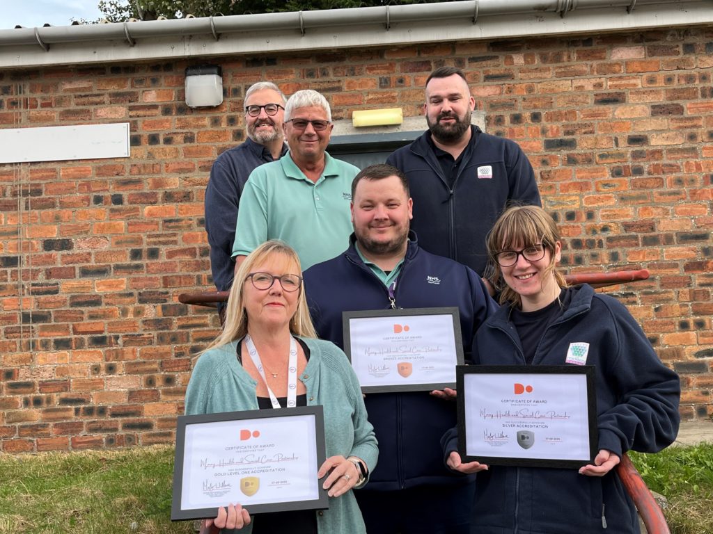Six members of staff from Health & Social Care Moray stand outside the Joint Equipment store building in Elgin and show the framed digital progress awards which have been achieved.