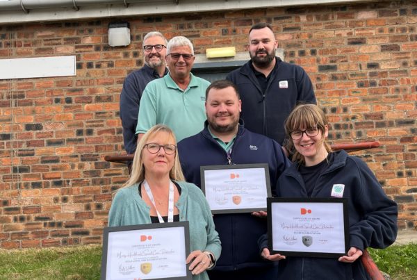Six members of staff from Health & Social Care Moray stand outside the Joint Equipment store building in Elgin and show the framed digital progress awards which have been achieved.