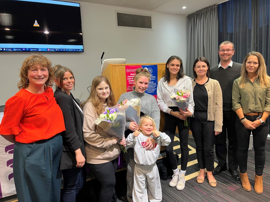 A group of adults and young adults, some holding bouquets of flowers, line up in front of a small stage in a conference room to mark the launch of the Maintaining relationships project.