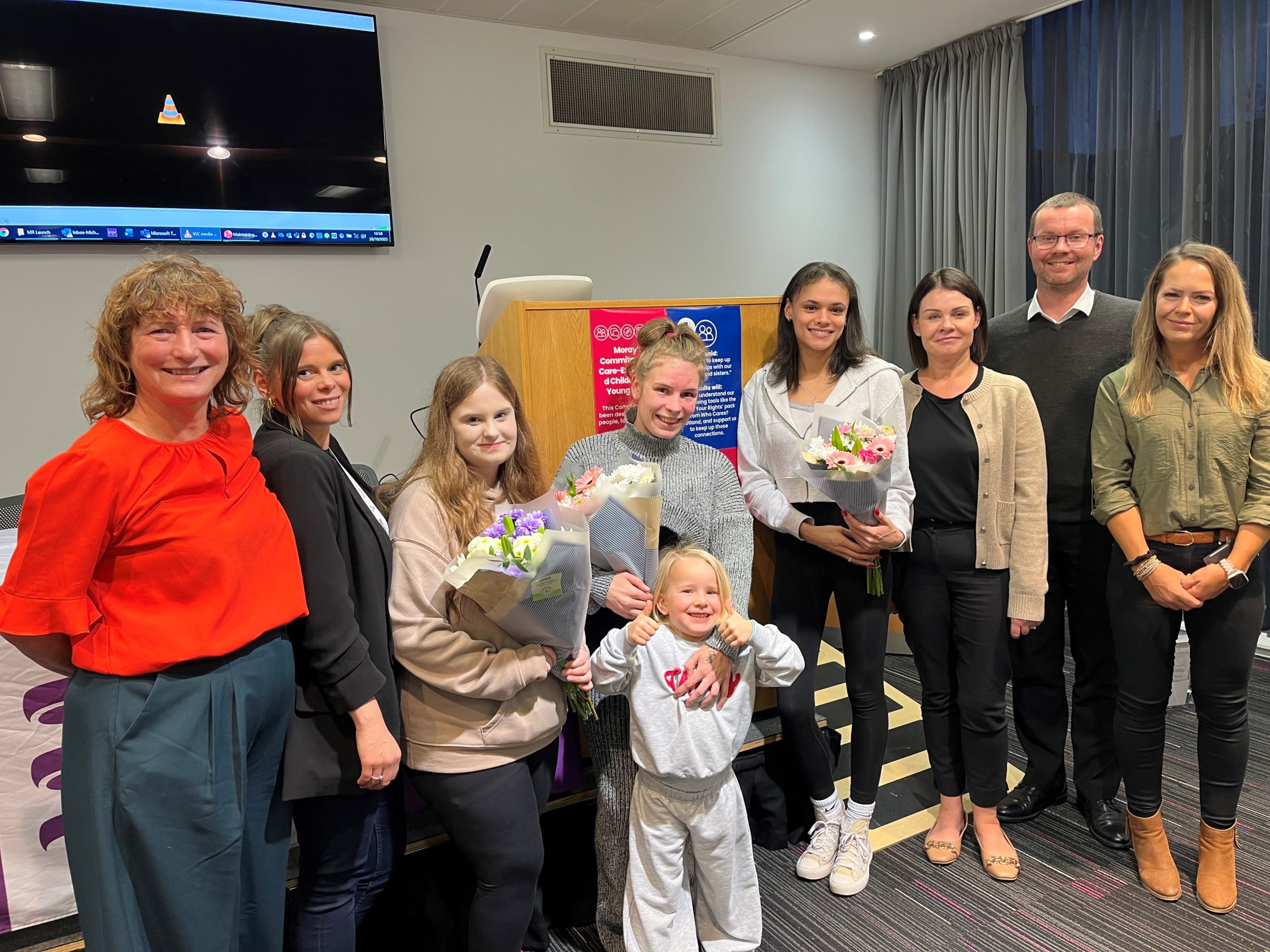 A group of adults and young adults, some holding bouquets of flowers, line up in front of a small stage in a conference room to mark the launch of the Maintaining relationships project.