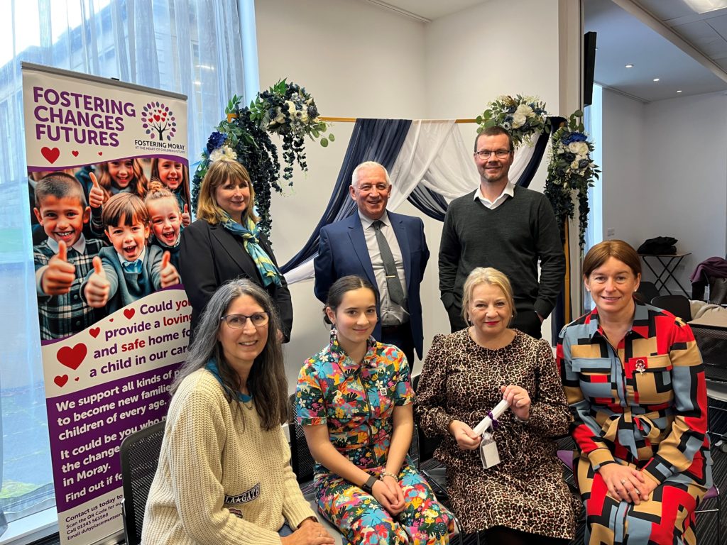 "A group of people pose indoors at a fostering event, standing in front of a decorative backdrop with draped fabric and flowers. A promotional banner on the left reads ‘Fostering Changes Futures – Fostering Moray with the Moray Council,’ encouraging people to provide safe homes for children. The individuals are dressed in business and casual attire, and one person holds a rolled-up certificate, suggesting a celebratory occasion."
