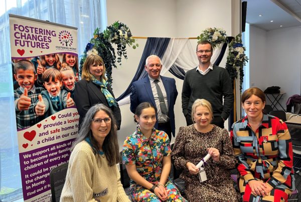 "A group of people pose indoors at a fostering event, standing in front of a decorative backdrop with draped fabric and flowers. A promotional banner on the left reads โFostering Changes Futures โ Fostering Moray with the Moray Council,โ encouraging people to provide safe homes for children. The individuals are dressed in business and casual attire, and one person holds a rolled-up certificate, suggesting a celebratory occasion."