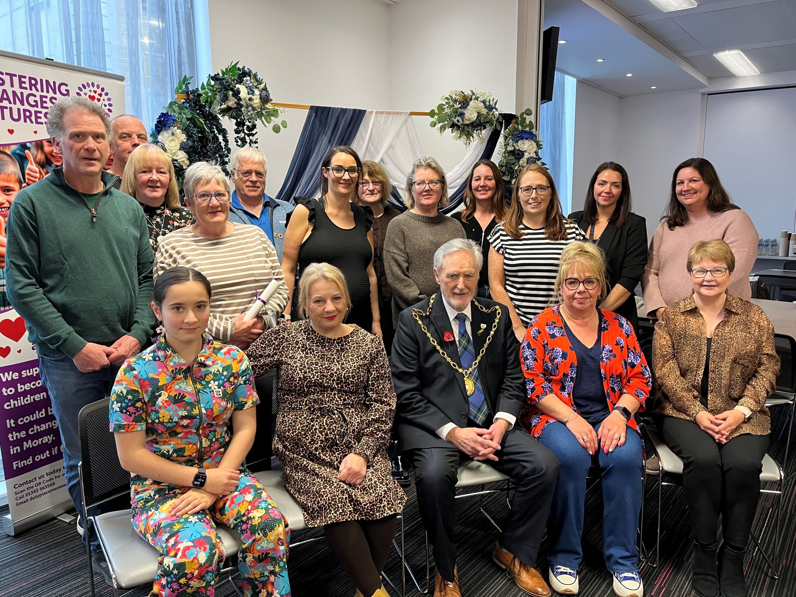 A mixed group of foster carers pose indoors at a fostering event, standing in front of a decorative backdrop with draped fabric and flowers. A promotional banner on the left reads ‘Fostering Changes Futures – Fostering Moray with the Moray Council,’ encouraging people to provide safe homes for children. Sitting front row centre is the civic leader of Moray Council. He is wearing a suit, tartan pattern tie and his gold chain of office.