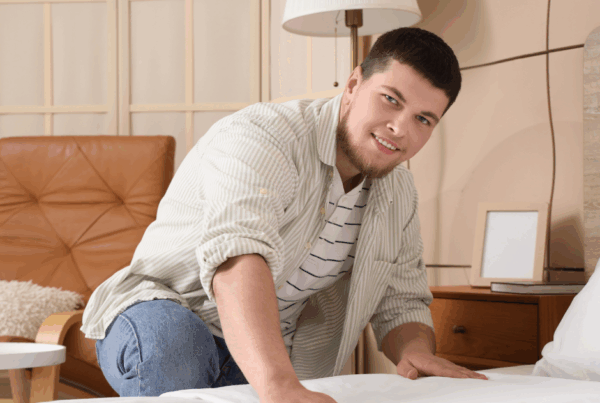 A happy and relaxed young man, dressed casually, kneels at the side of his nearly made bed in his tidy bedroom, smoothing down the sheets.