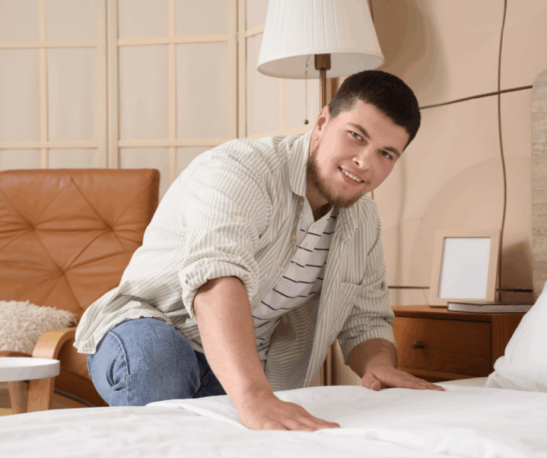 A happy and relaxed young man, dressed casually, kneels at the side of his nearly made bed in his tidy bedroom, smoothing down the sheets.
