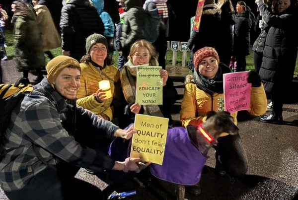 A group of people, out on a dark night and wrapped up against the cold with thick jackets and hats, holding colourful printed posters calling for an end to gender violence.