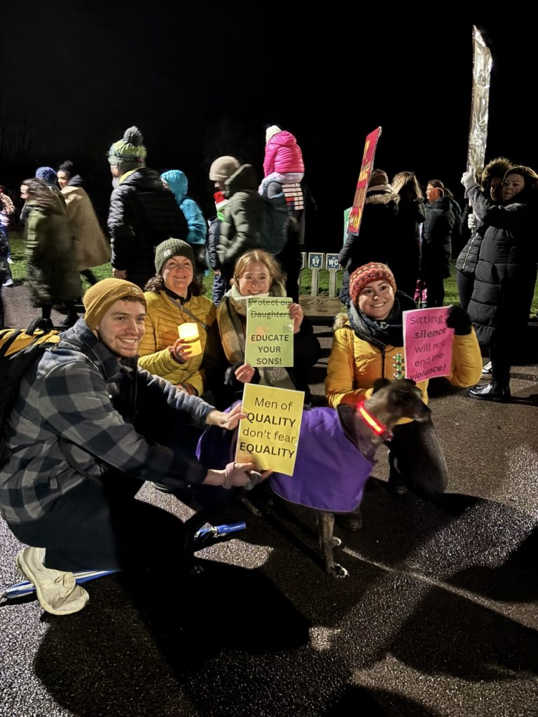 A group of people, out on a dark night and wrapped up against the cold with thick jackets and hats, holding colourful printed posters calling for an end to gender violence.