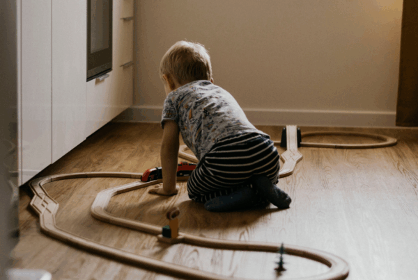 A child sits on the floor of a bedroom, playing with a wooden train set.
