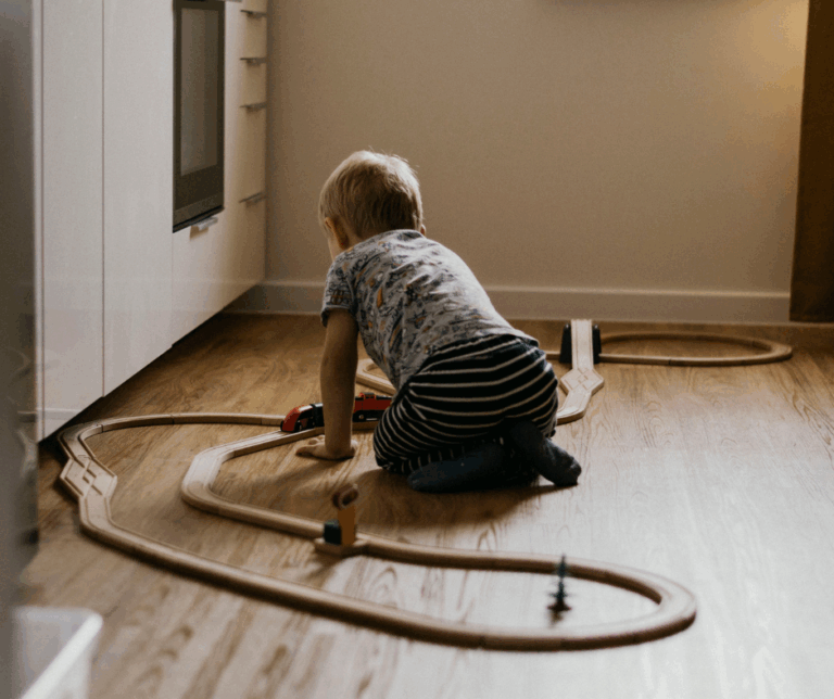 A child sits on the floor of a bedroom, playing with a wooden train set.