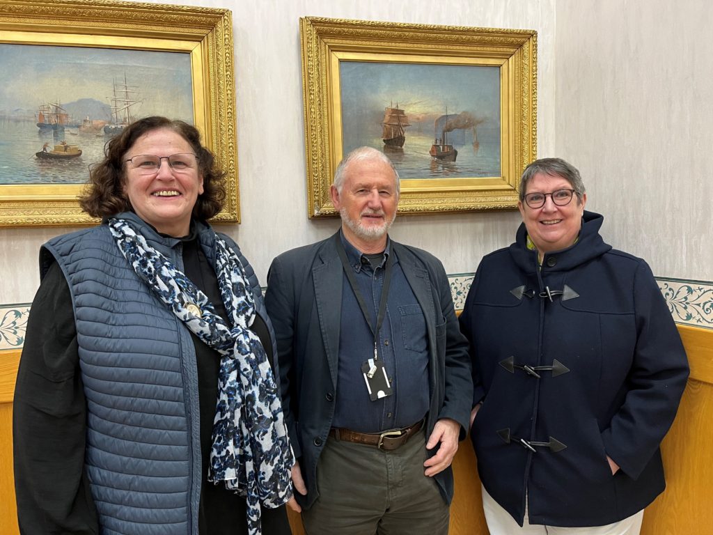 Three people standing together indoors, smiling at the camera. They are new carer representatives Miriam Connor and Cjristine Stevens, with outgoing member Ivan Augustus standing in the middle. Behind them are two ornate gold-framed paintings depicting sailing ships on water."