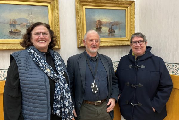 Three people standing together indoors, smiling at the camera. They are new carer representatives Miriam Connor and Cjristine Stevens, with outgoing member Ivan Augustus standing in the middle. Behind them are two ornate gold-framed paintings depicting sailing ships on water."