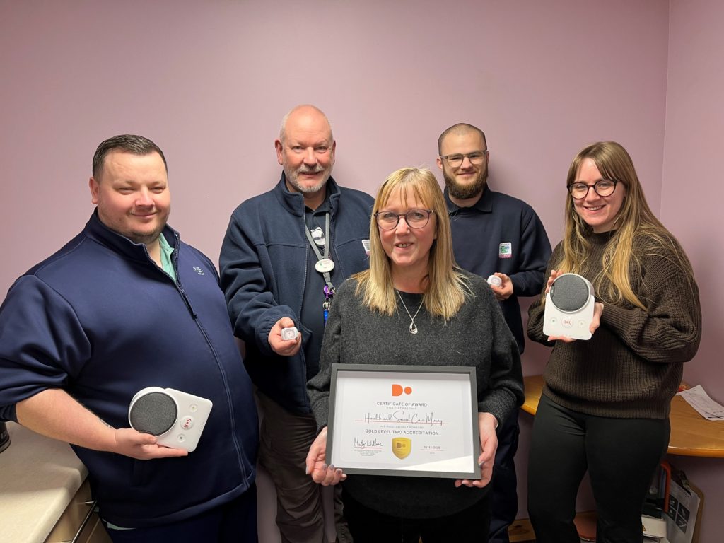 A mixed group of five HSCM staff members gather in an office to proudly display a framed certificate. Some are holding up telecare equipment.