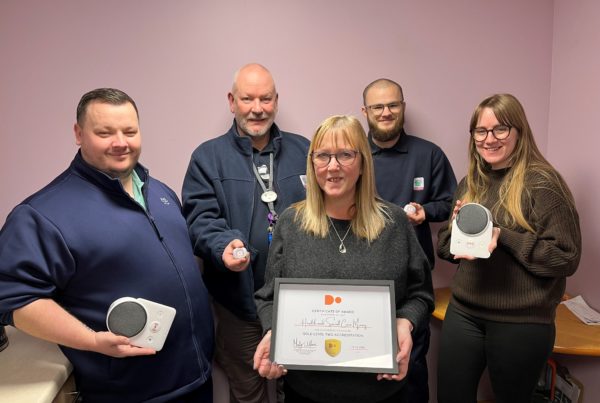 A mixed group of five HSCM staff members gather in an office to proudly display a framed certificate. Some are holding up telecare equipment.