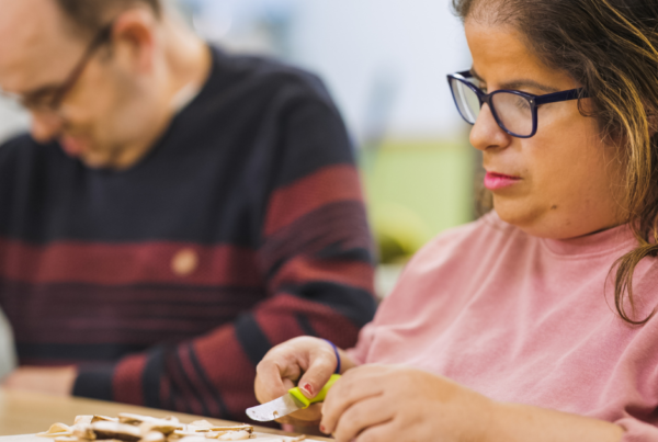 Two adults sitting working at a count top. The man in the background has glasses and is wearing a stripy jumper. The woman who is closer to the camera also wear glasses, has shoulder length hair and is wearing a pink top. She is working at a chopping board and is preparing mushrooms with a knife.