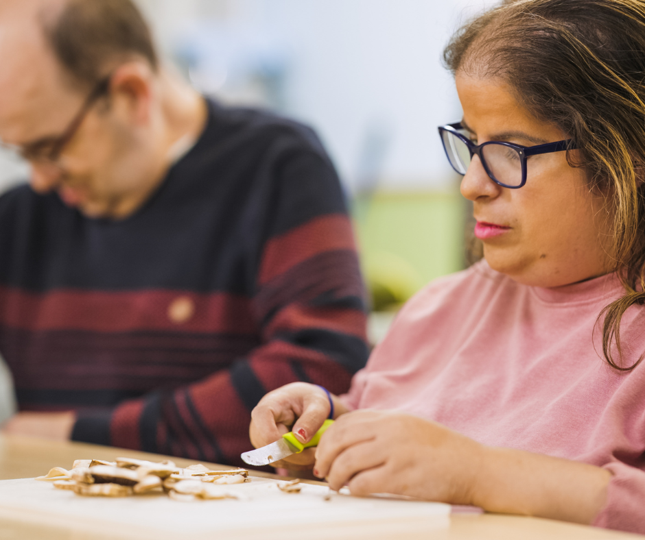 Two adults sitting working at a count top. The man in the background has glasses and is wearing a stripy jumper. The woman who is closer to the camera also wear glasses, has shoulder length hair and is wearing a pink top. She is working at a chopping board and is preparing mushrooms with a knife.