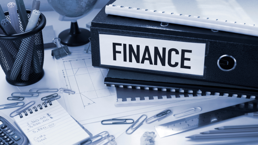 A close-up view of a busy desk in an office with a calculator, notebooks, paperclips, pencils, and a container with pens. There are some official looking bound files on the desk, including a box file which has the word finance printed on it.