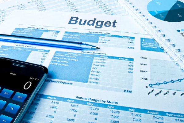 A budget planning spreadsheet showing monthly income, expenses, and planned expenditures, alongside a calculator, pen, pie chart, and line graph, all in a blue colour scheme.