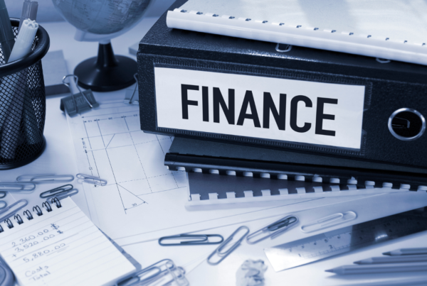 A close-up view of a busy desk in an office with a calculator, notebooks, paperclips, pencils, and a container with pens. There are some official looking bound files on the desk, including a box file which has the word finance printed on it.