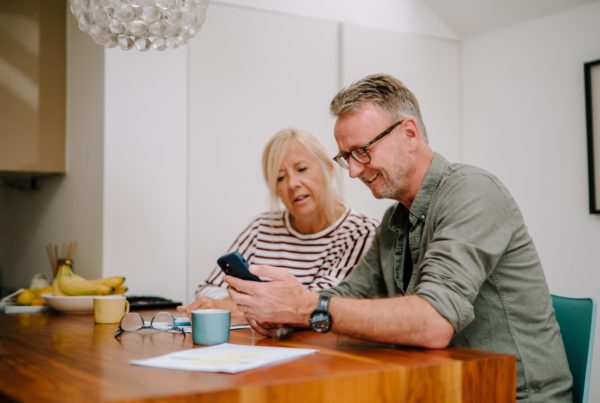 Two middle aged people, a man and a woman, sit together at a wooden table in a home. Both are dressed in everyday clothes. They are looking at the screen of a mobile phone being held by the man. There are mugs, papers and a pair of reading glasses on the table.