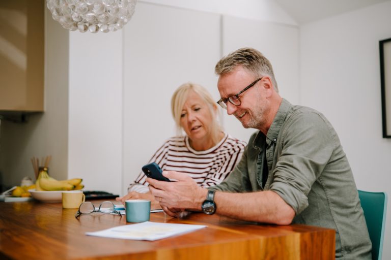 Two middle aged people, a man and a woman, sit together at a wooden table in a home. Both are dressed in everyday clothes. They are looking at the screen of a mobile phone being held by the man. There are mugs, papers and a pair of reading glasses on the table.