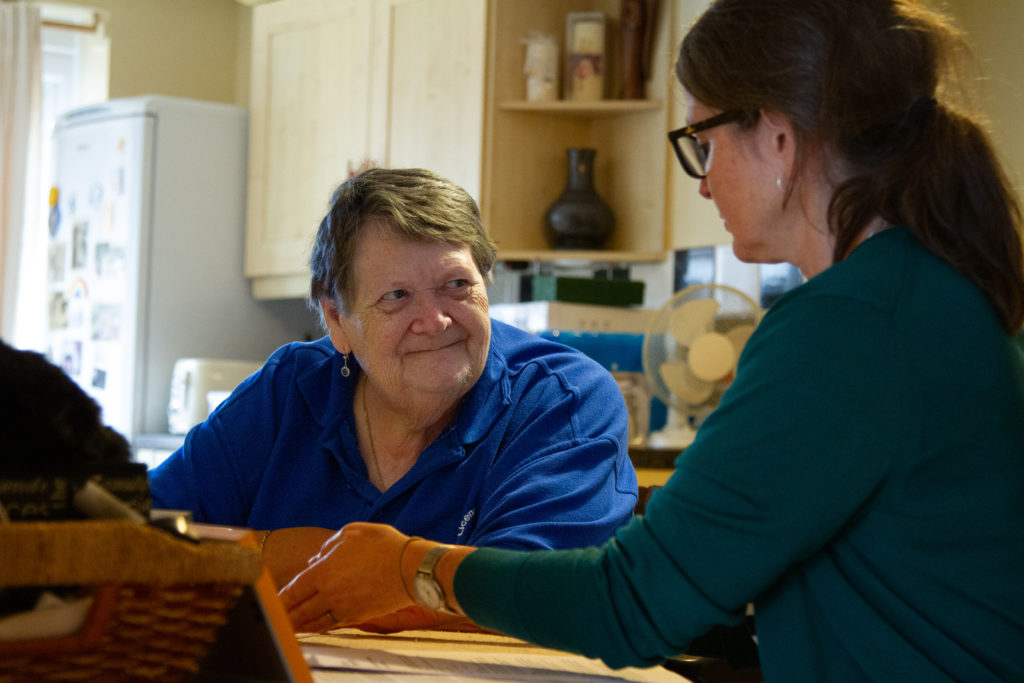 Two women sit together relaxed and talking at a table in the kitchen of a home. One is front on to the camera and the other is in side profile.