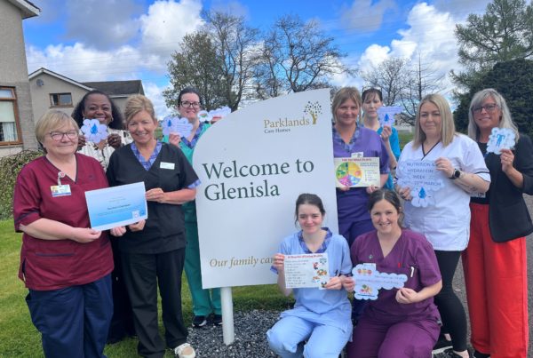 A group of care staff stand and kneel outdoors around a sign reading ‘Welcome to Glenisla’ at Parklands Care Homes. The staff wear colourful uniforms and hold posters about nutrition and hydration awareness, with a care home building, trees, and blue sky in the background.”