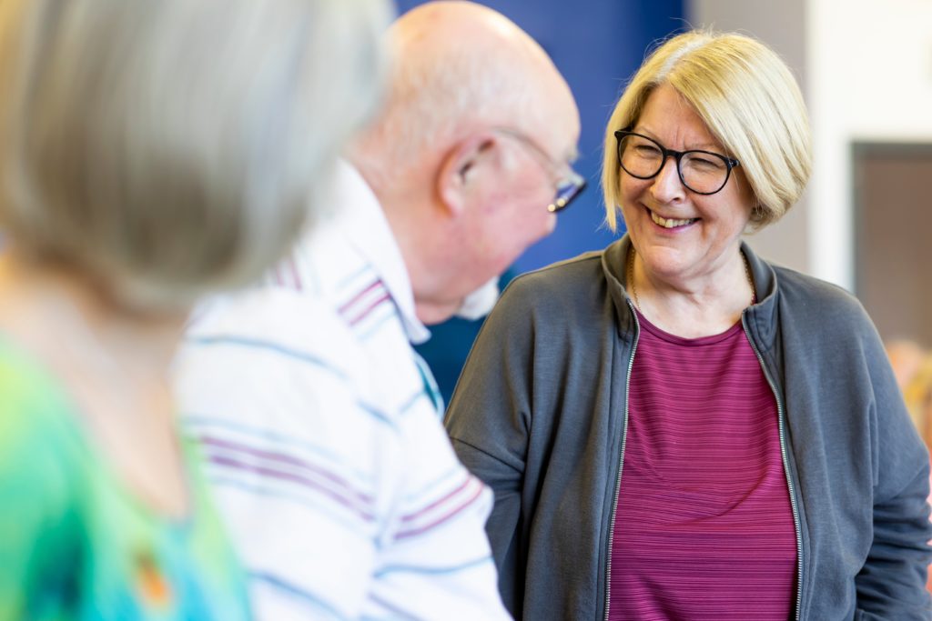 Two older adults stand and face each other in a bright indoor setting, one in sharp focus wearing glasses and a grey jacket over a pink top, while another balding person in a striped shirt is slightly out of focus in the foreground; a third blurred figure appears at the left edge of the frame.