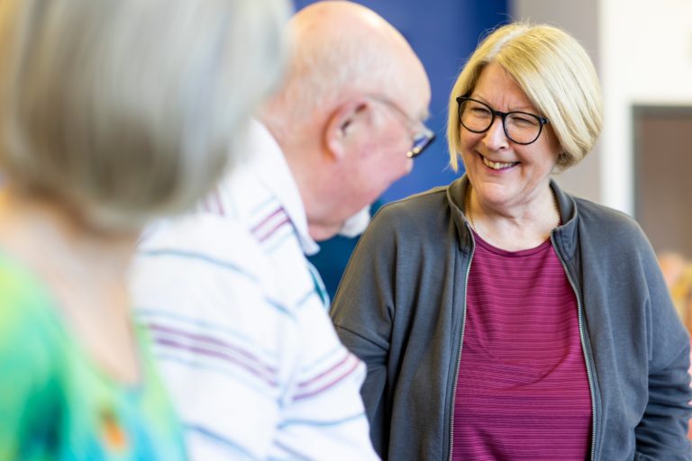 Two older adults stand and face each other in a bright indoor setting, one in sharp focus wearing glasses and a grey jacket over a pink top, while another balding person in a striped shirt is slightly out of focus in the foreground; a third blurred figure appears at the left edge of the frame.