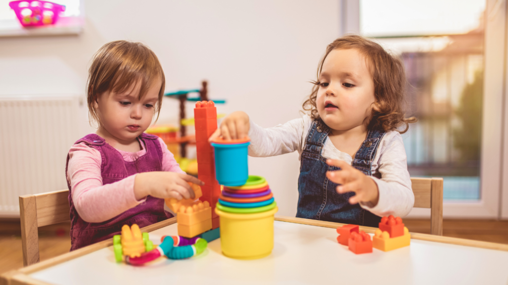 Two toddlers sit together at a table. They are playing with colourful toys including cups and shapes.