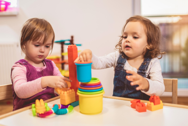 Two toddlers sit together at a table. They are playing with colourful toys including cups and shapes.