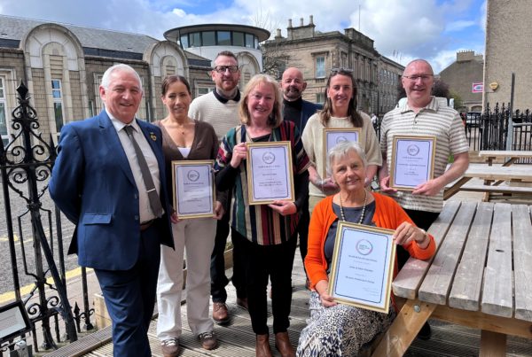 Group of eight adults posing outdoors in a courtyard, several holding framed certificates labeled “Health & Social Care Moray,” while one person sits at a wooden picnic table in front also holding an award.
