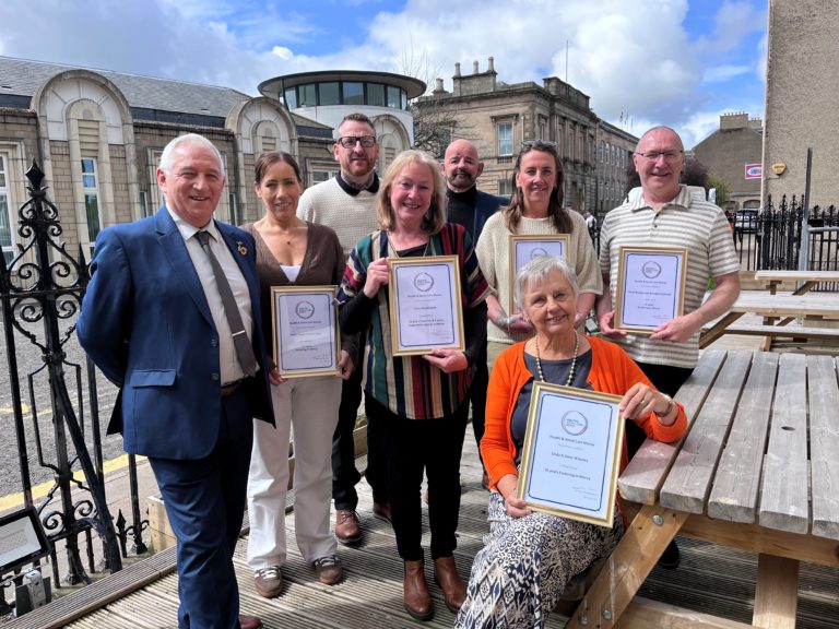 Group of eight adults posing outdoors in a courtyard, several holding framed certificates labeled “Health & Social Care Moray,” while one person sits at a wooden picnic table in front also holding an award.