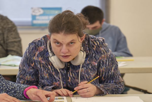 A woman with over-ear headphones sits at a long table, holding a pencil and working on a worksheet. There is another row of people in the background.