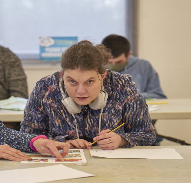 A woman with over-ear headphones sits at a long table, holding a pencil and working on a worksheet. There is another row of people in the background.