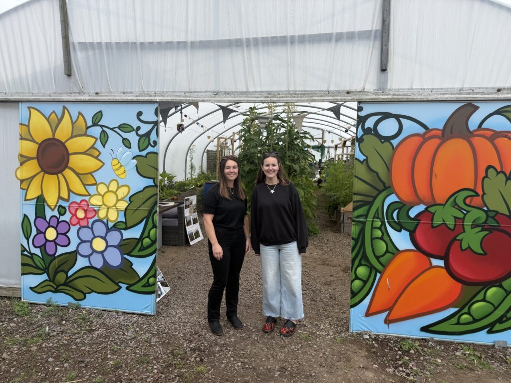 Two people stand at the entrance of a polytunnel garden. Colourful painted panels on either side feature flowers, a bee, a pumpkin, tomatoes and other vegetables. Raised beds and tall plants are visible inside the polytunnel behind them.