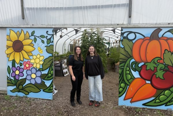 Two people stand at the entrance of a polytunnel garden. Colourful painted panels on either side feature flowers, a bee, a pumpkin, tomatoes and other vegetables. Raised beds and tall plants are visible inside the polytunnel behind them.