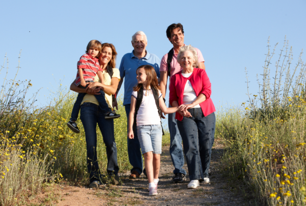 Four adults and two children from a multi-generation family walk together along a sunlit countryside path bordered by tall grasses and yellow wildflowers beneath a clear blue sky.