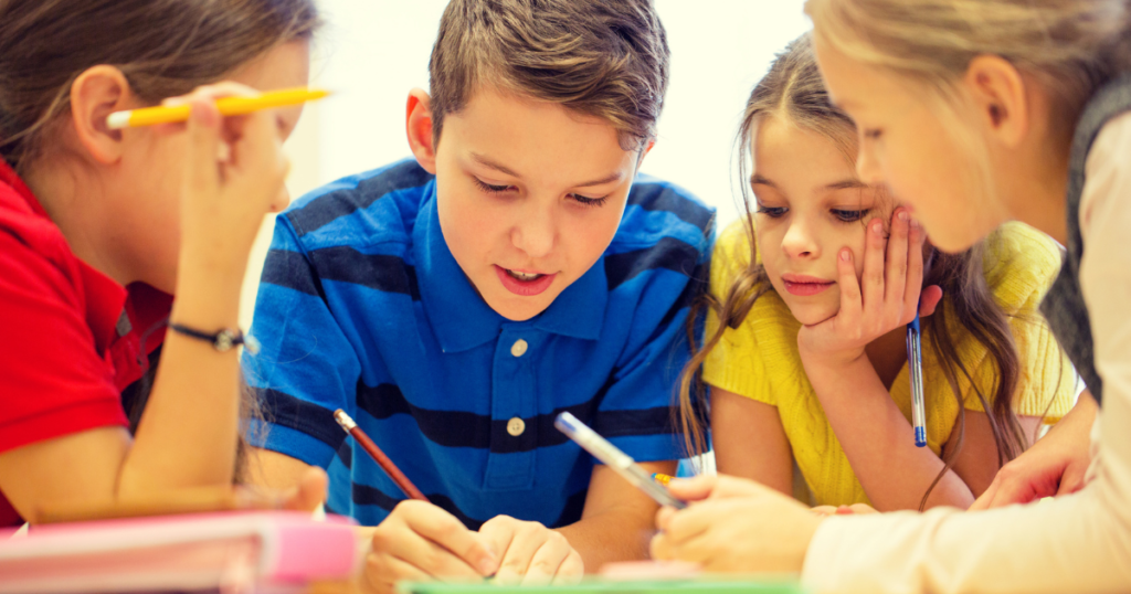 Close-up of four children gathered around a table, writing and discussing a shared piece of work. Pens, pencils and study materials are visible as the group collaborates on a classroom activity.