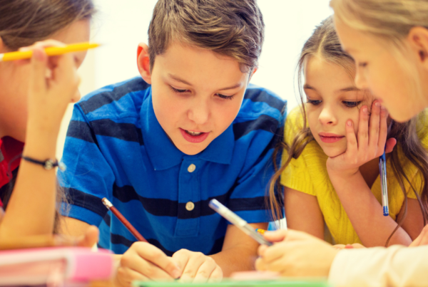 Close-up of four children gathered around a table, writing and discussing a shared piece of work. Pens, pencils and study materials are visible as the group collaborates on a classroom activity.