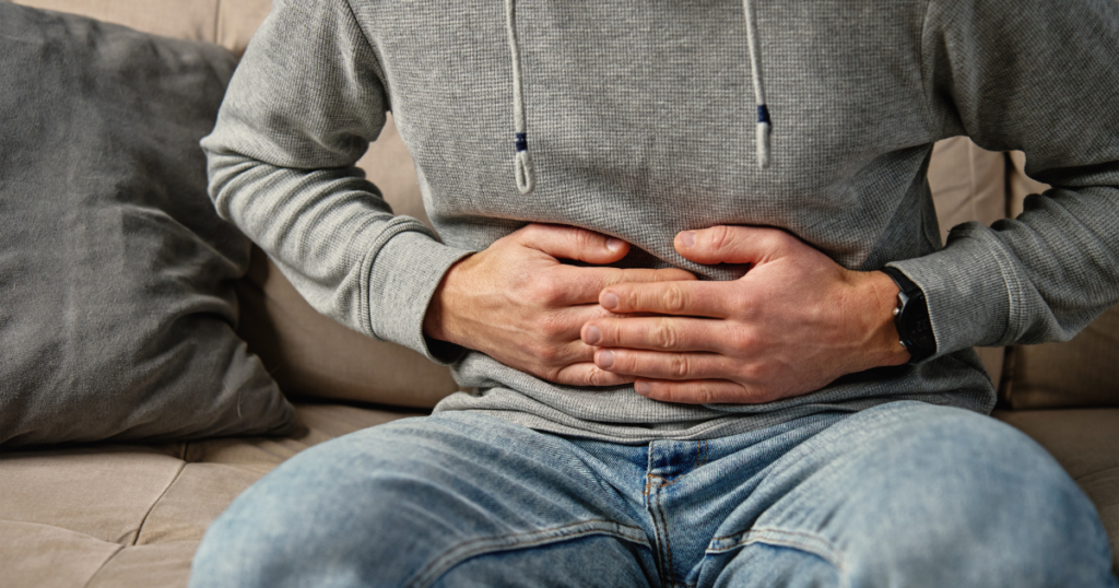 Close-up of a person sitting on a sofa with both hands pressed against their abdomen. The person is wearing a grey hooded sweatshirt, blue jeans and a wristwatch. A cushion is visible beside them, and the image appears to represent stomach pain, digestive discomfort or abdominal health concerns.