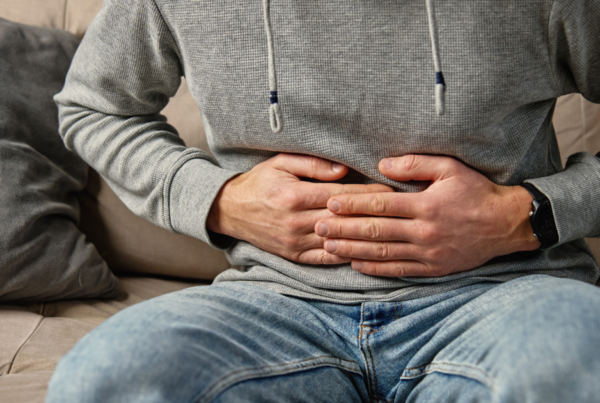 Close-up of a person sitting on a sofa with both hands pressed against their abdomen. The person is wearing a grey hooded sweatshirt, blue jeans and a wristwatch. A cushion is visible beside them, and the image appears to represent stomach pain, digestive discomfort or abdominal health concerns.