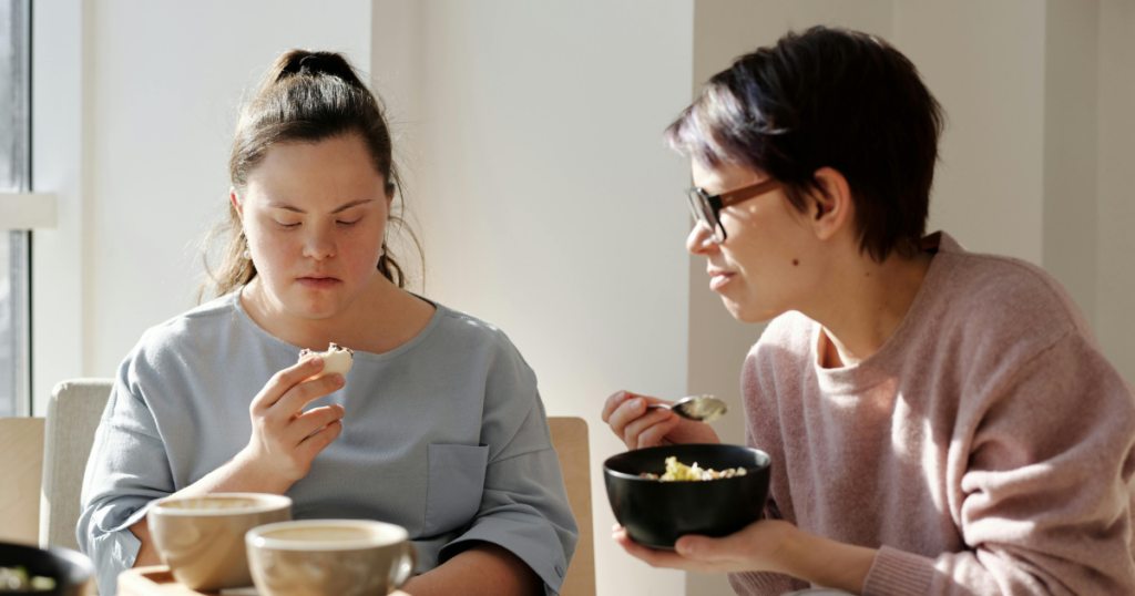 Two people sit at a table in a bright, sunlit room. One person in a light blue top is holding a small piece of food and looking down at it, while another person wearing glasses and a pale pink jumper holds a bowl and spoon. Two cups sit on the table in the foreground, and natural light streams in from a nearby window.