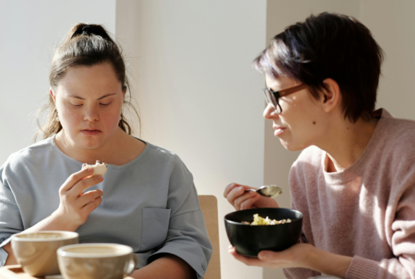Two people sit at a table in a bright, sunlit room. One person in a light blue top is holding a small piece of food and looking down at it, while another person wearing glasses and a pale pink jumper holds a bowl and spoon. Two cups sit on the table in the foreground, and natural light streams in from a nearby window.