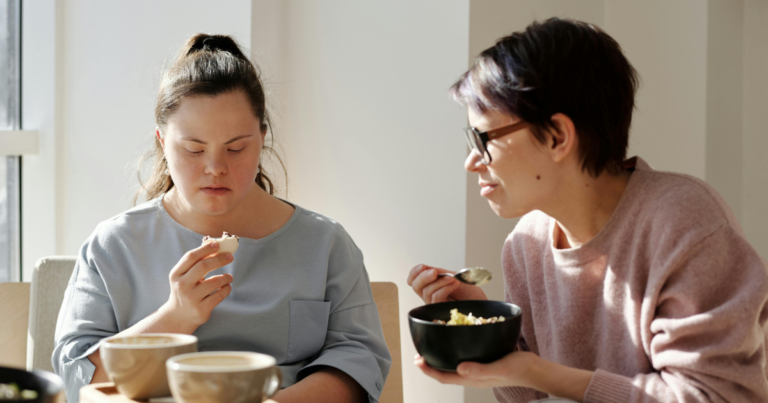 Two people sit at a table in a bright, sunlit room. One person in a light blue top is holding a small piece of food and looking down at it, while another person wearing glasses and a pale pink jumper holds a bowl and spoon. Two cups sit on the table in the foreground, and natural light streams in from a nearby window.