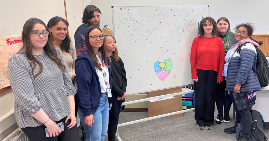 A group of young people and adults stand around a whiteboard in a classroom or meeting room. The whiteboard is decorated with colourful confetti-style markings and reads “Welcome to Moray” with a multicoloured heart drawn underneath. Posters are displayed on the walls, and the group faces the camera beside the welcome display.